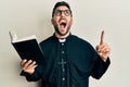 Young hispanic priest man holding bible with finger up angry and mad screaming frustrated and furious, shouting with anger looking