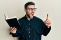 Young hispanic priest man holding bible with finger up angry and mad screaming frustrated and furious, shouting with anger