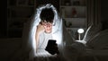 Young hispanic teenager seriously engrossed in texting on his smartphone, lying on a comfy bed under a snug blanket, in the dim Royalty Free Stock Photo