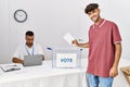 Young hispanic voter man smiling happy putting vote in ballot box at electoral center