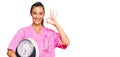 Young hispanic woman as nutritionist doctor holding weighing machine doing ok sign with fingers, smiling friendly gesturing