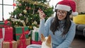 Young hispanic woman with chihuahua dog listening to music wearing headphones and christmas hat at home