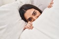 Young hispanic woman covering face with bedsheet lying on bed at bedroom