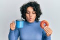 Young hispanic woman with curly hair drinking a cup of coffee and croissant making fish face with mouth and squinting eyes, crazy