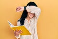 Young hispanic woman with curly hair holding book smiling cheerful playing peek a boo with hands showing face