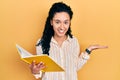 Young hispanic woman with curly hair holding book smiling cheerful presenting and pointing with palm of hand looking at the camera Royalty Free Stock Photo