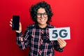 Young hispanic woman with curly hair holding 5g technology smartphone winking looking at the camera with sexy expression, cheerful Royalty Free Stock Photo