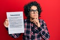 Young hispanic woman with curly hair holding insurance document serious face thinking about question with hand on chin, thoughtful Royalty Free Stock Photo