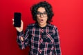 Young hispanic woman with curly hair holding smartphone showing blank screen scared and amazed with open mouth for surprise, Royalty Free Stock Photo