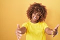 Young hispanic woman with curly hair standing over yellow background looking at the camera smiling with open arms for hug Royalty Free Stock Photo