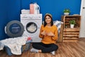 Young hispanic woman doing laundry hands together and fingers crossed smiling relaxed and cheerful