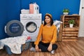 Young hispanic woman doing laundry smiling looking to the side and staring away thinking