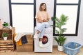 Young hispanic woman drinking coffee waiting for washing machine at laundry room looking to side, relax profile pose with natural