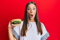 Young hispanic woman eating hamburger scared and amazed with open mouth for surprise, disbelief face