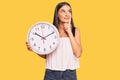 Young hispanic woman holding big clock serious face thinking about question with hand on chin, thoughtful about confusing idea Royalty Free Stock Photo