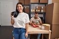 Young hispanic woman holding camera working at small business ecommerce looking positive and happy standing and smiling with a Royalty Free Stock Photo