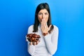 Young hispanic woman holding dates bowl covering mouth with hand, shocked and afraid for mistake