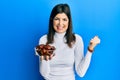 Young hispanic woman holding dates bowl screaming proud, celebrating victory and success very excited with raised arm