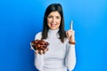 Young hispanic woman holding dates bowl smiling with an idea or question pointing finger with happy face, number one