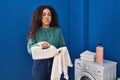 Young hispanic woman holding dirty laundry and detergent bottle puffing cheeks with funny face Royalty Free Stock Photo