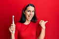 Young hispanic woman holding electric toothbrush pointing thumb up to the side smiling happy with open mouth