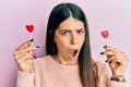 Young hispanic woman holding heart lollipops in shock face, looking skeptical and sarcastic, surprised with open mouth Royalty Free Stock Photo