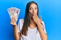 Young hispanic woman holding japanese yen banknotes covering mouth with hand, shocked and afraid for mistake