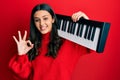 Young hispanic woman holding piano keyboard doing ok sign with fingers, smiling friendly gesturing excellent symbol