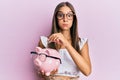 Young hispanic woman holding piggy bank with glasses and coin puffing cheeks with funny face