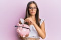 Young hispanic woman holding piggy bank with glasses and coin relaxed with serious expression on face Royalty Free Stock Photo
