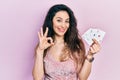 Young hispanic woman holding poker cards doing ok sign with fingers, smiling friendly gesturing excellent symbol