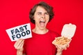 Young hispanic woman holding potato chip and fast food banner angry and mad screaming frustrated and furious, shouting with anger