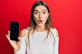Young hispanic woman holding smartphone showing blank screen scared and amazed with open mouth for surprise, disbelief face Royalty Free Stock Photo