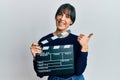 Young hispanic woman holding video film clapboard pointing thumb up to the side smiling happy with open mouth