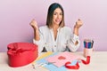 Young hispanic woman making valentine gift sitting on the table very happy and excited doing winner gesture with arms raised, Royalty Free Stock Photo