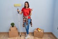 Young hispanic woman painting home walls with paint roller with a big smile on face, pointing with hand finger to the side looking Royalty Free Stock Photo