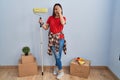 Young hispanic woman painting home walls with paint roller covering one eye with hand, confident smile on face and surprise Royalty Free Stock Photo