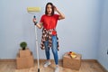 Young hispanic woman painting home walls with paint roller crazy and scared with hands on head, afraid and surprised of shock with Royalty Free Stock Photo