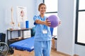 Young hispanic woman physiotherapist stretching arms using ball at rehab clinic