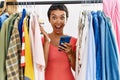 Young hispanic woman with short hair searching clothes on clothing rack using smartphone celebrating victory with happy smile and Royalty Free Stock Photo