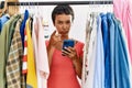 Young hispanic woman with short hair searching clothes on clothing rack using smartphone pointing with finger to the camera and to Royalty Free Stock Photo