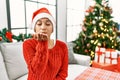 Young hispanic woman with short hair wearing christmas hat sitting on the sofa looking at the camera blowing a kiss with hand on Royalty Free Stock Photo
