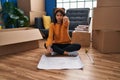 Young hispanic woman sitting on the floor at new home looking at blueprints pointing with finger to the camera and to you, Royalty Free Stock Photo