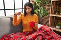 Young hispanic woman sitting on the sofa drinking a coffee at home pointing down looking sad and upset, indicating direction with Royalty Free Stock Photo