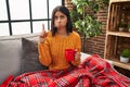 Young hispanic woman sitting on the sofa drinking a coffee at home pointing up looking sad and upset, indicating direction with Royalty Free Stock Photo