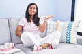 Young hispanic woman sitting on the sofa at home smiling cheerful presenting and pointing with palm of hand looking at the camera Royalty Free Stock Photo