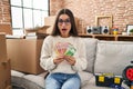 Young hispanic woman sitting on the sofa at new home holding money afraid and shocked with surprise and amazed expression, fear Royalty Free Stock Photo