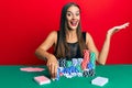 Young hispanic woman sitting on the table holding casino chips celebrating achievement with happy smile and winner expression with Royalty Free Stock Photo