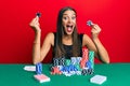 Young hispanic woman sitting on the table holding casino chips celebrating crazy and amazed for success with open eyes screaming Royalty Free Stock Photo