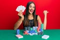 Young hispanic woman sitting on the table playing poker holding cards pointing thumb up to the side smiling happy with open mouth